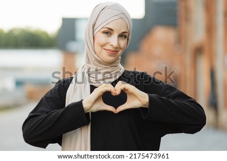 Beautiful young muslim woman smiling confident, enthusiastic and cheerful with hands sign of love, affection, happy, on chest walking on the background of city.