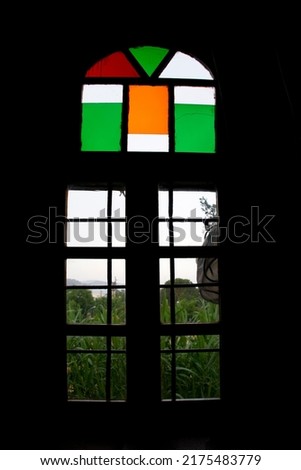 A silhouette image looking through an arched window in a Greek Orthodox church to green fields beyond.  Stained glass panels are at the top in green, orange and red .