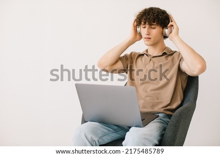 Attractive young man using laptop, wearing headphones while sitting in armchair against white wall, copy space. Millennial male communicating online, working or studying remotely on portable pc