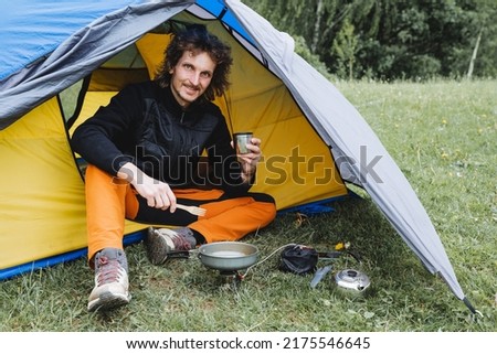 A curly-haired hiker hiking guy has lunch in a tent, a male camper sitting in a campsite cooking food on a gas burner, equipment for relaxing in the forest. High quality photo