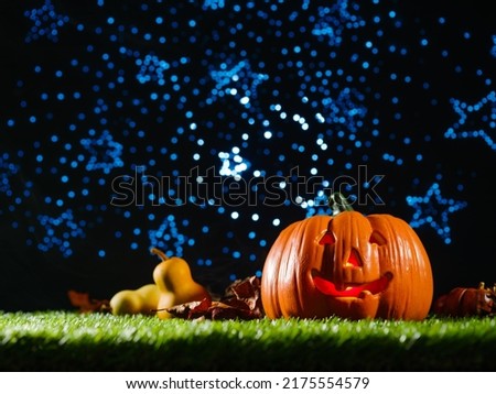 Halloween composition on the background of a blue starry sky. Orange pumpkin with carved face and smile, autumn fruits and cones on green grass. There are no people in the photo.