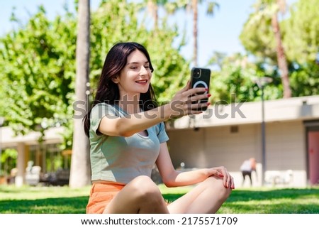 Young woman smiling confident wearing turquoise tee and orange short on city park, outdoors taking a self portrait with smart phone. She looks at the screen and taking selfie.
