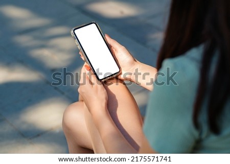 Beautiful brunette woman wearing turquoise t-shirt standing on city park, outdoors hands holding phone touching finger mockup white blank display, mobile app tech concept, over shoulder closeup view. Royalty-Free Stock Photo #2175571741