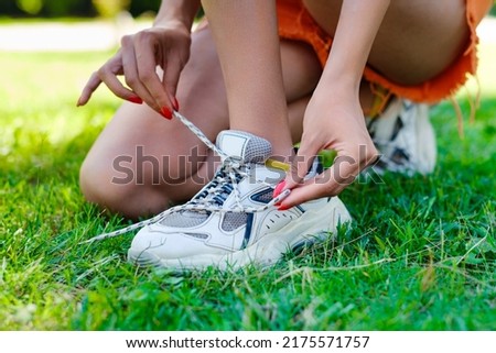 Young brunette woman wearing turquoise tee and orange short on city park, outdoors tying lace running shoes getting ready for run. Jogging girl exercise motivation health and fitness.