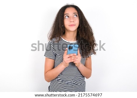 Portrait of young beautiful brunette woman wearing striped t-shirt over white wall with dreamy look, thinking while holding smartphone. Tries to write up a message.