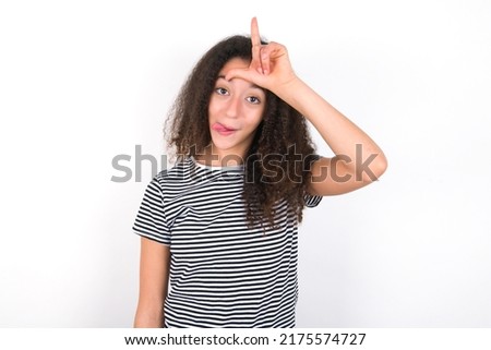 young beautiful brunette woman wearing striped t-shirt over white wall gestures with finger on forehead makes loser gesture makes fun of people shows tongue