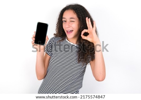 Excited young beautiful brunette woman wearing striped t-shirt over white wall showing smartphone blank screen, blinking eye and doing ok sign with hand.  Advertisement concept.