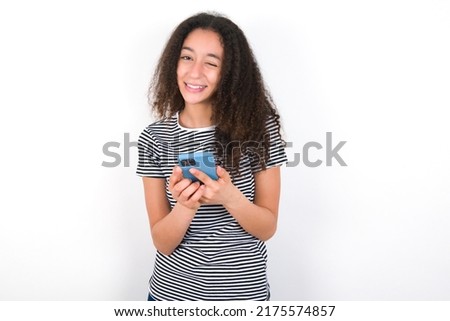 Pleased young beautiful brunette woman wearing striped t-shirt over white wall using self phone and looking and winking at the camera. Flirt and coquettish concept.