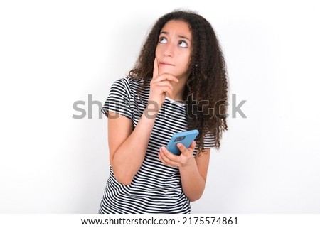 Image of a thinking dreaming young beautiful brunette woman wearing striped t-shirt over white wall using mobile phone and holding hand on face. Taking decisions and social media concept.