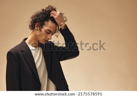 Defiant awesome tanned curly man in classic brown jacket touches curls posing isolated on over beige pastel background. Fashion New Collection offer. Retro style concept. Free place for ad
