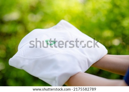 Hand holding a white shirt made from recycled materials. Recycle symbol on the shirt with green blurred background.