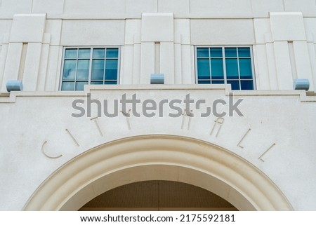 The exterior facade of a vintage city hall building with white limestone walls closed glass windows reflecting the sky and yellow trim. The words city hall is spelled on the doorway arch in thin font
