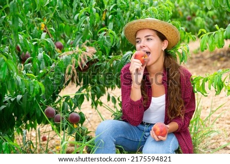 Young lady eating juicy peach while sitting amongst trees in plantation. Royalty-Free Stock Photo #2175594821