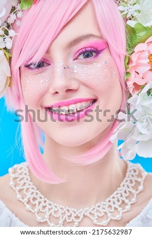 Beauty, makeup and hairstyle. Portrait of a pretty, smiling teen girl with bright pink make-up posing in colored pink wig and flower wreath on head. Studio portrait on a blue background.
