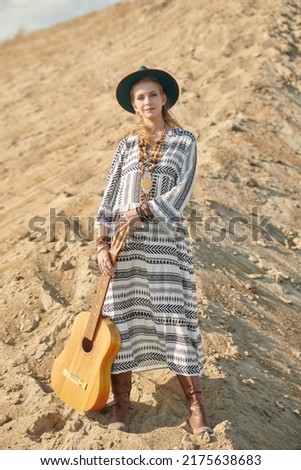 A girl in an ethnic boho dress and hat walks along the crest of a sand dune with a guitar. Spirit of freedom, search for adventure.