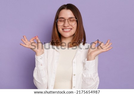 Image of positive satisfied delighted young adult Caucasian woman wearing white shirt, practicing yoga, relaxing, keeps eyes closed, posing isolated over purple background.