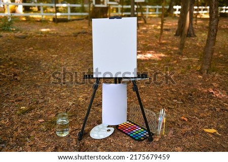 Canvas place with empty white board with a lot of painting tools like paint, brushes and palette. Invitation for people to paint their message in a garden.