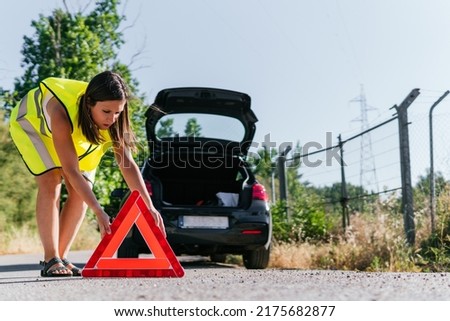 Woman with yellow reflective vest placing the emergency warning system for her broken down car. Young girl supporting a warning triangle for broken down vehicles. Royalty-Free Stock Photo #2175682877
