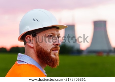 Portrait of a nuclear power plant engineer. A man with a beard in a protective helmet and an orange vest stands against the background of a nuclear power plant Royalty-Free Stock Photo #2175706851