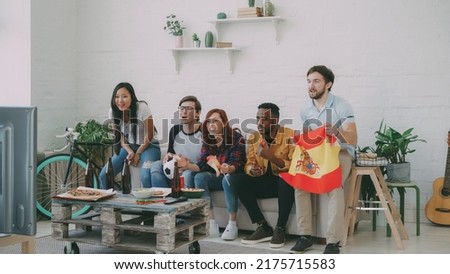 Multi-ethnic group of friends sports fans with Spainsh flags watching football championship on TV together at home indoors and cheering up favourite team Royalty-Free Stock Photo #2175715583