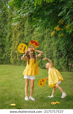 Two amusing girls with a variety of cardboard emoticons in their hands are having fun in the park on the lawn. Children indulge and make faces