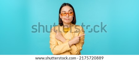 Indecisive asian woman in sunglasses, makes choice between two variants, points sideways, looks puzzled, stands over blue background Royalty-Free Stock Photo #2175720889
