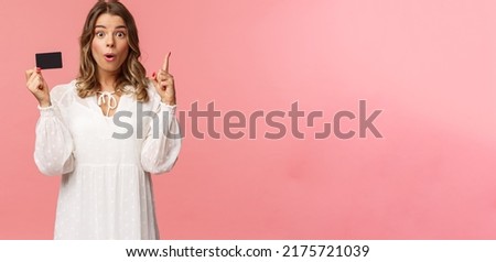 Portrait of thoughtful cute blond girl in white dress, have and excellent idea, holding credit card say wow, eureka, raise index finger have suggestion, know where find needed product