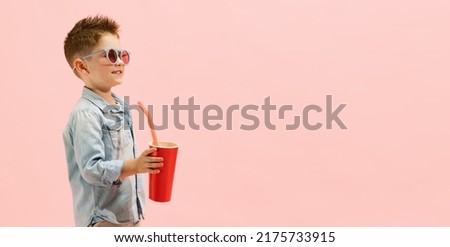 Portrait of stylish little boy, child in jeans t-shirt and sunglasses, drinking lemonade isolated over pink background. Summertime mood. Concept of childhood, family, emotions, lifestyle, fashion, ad