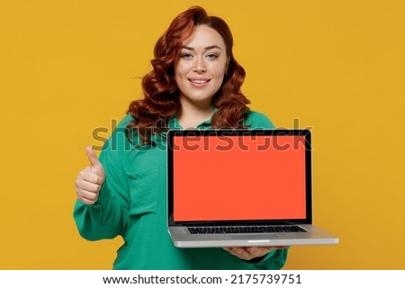 Young ginger chubby overweight woman 20s wears green shirt hold use work on laptop pc computer with blank screen workspace area show thumb up like isolated on plain yellow background studio portrait