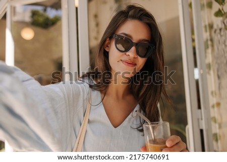 Cute young caucasian woman wears casual summer clothes, sunglasses taking selfie outdoors. Brunette is holding glass with cool drink. Lifestyle concept