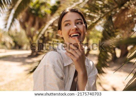 Cheerful young caucasian woman looking away smiling with her teeth standing in tropical garden. Brunette with collected hair wears white shirt. Positive emotions concept