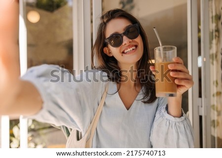 Beautiful young caucasian woman with chocolate smoothie takes selfie outdoors. Brunette wears sunglasses and blue dress in summer. Lifestyle concept