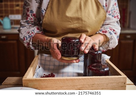 Cropped view of a woman in kitchen apron holding a glass jar of redcurrant jam over a wooden box with berry marmalade jars upside down. Collection of homemade preserves, confitures, jelly, canned food Royalty-Free Stock Photo #2175746351