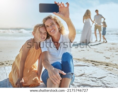 Cheerful mature woman and little girl taking a selfie while sitting on the beach. Happy little girl smiling while sitting with her mom or grandmother taking picture on mobile phone while on vacation