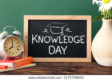 Vase with flowers, stationery and chalkboard with text KNOWLEDGE DAY on wooden table