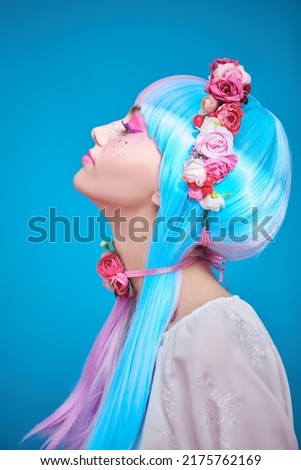 Sideview portrait of a pretty teen girl with bright pink make-up posing in colored violet-blue wig and flower wreath on the head. Studio portrait on a blue background. Beauty, makeup and hairstyle. 