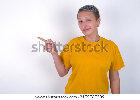 Caucasian woman with short hair wearing yellow T-shirt over white wall points to side on blank space demonstrates advertisement. People and promotion concept