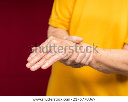 Senior woman suffering from wrist pain while standing against a red background. Close-up photo. Aged people and healthcare concept