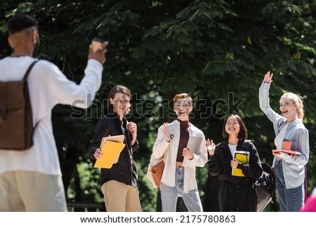 Positive interracial students waving hands near blurred african american friend in park Royalty-Free Stock Photo #2175780863