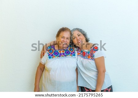 Portrait of two Mexican women smiling, mature daughter hugging her elderly mother, standing on white background, looking at camera. Wearing indigenous clothing with colorful flower print blouses
