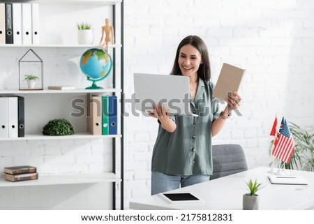 Positive teacher holding book and laptop during online lesson in school