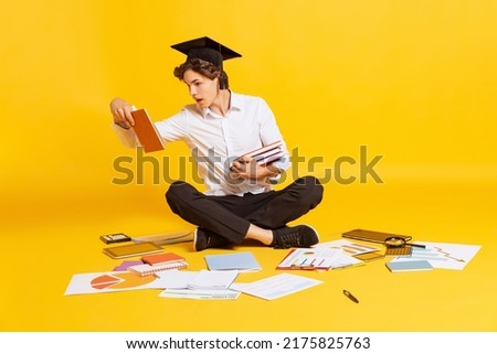 Shock. Young boy, student in business style clothes sitting on floor among books and notebooks isolated on yellow background. Exam preparation. Concept of education, studying, youth, back to school Royalty-Free Stock Photo #2175825763