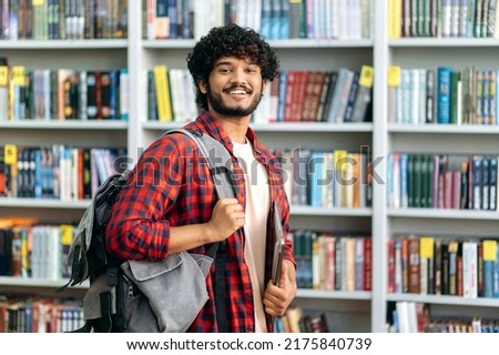 Handsome proud curly haired indian or arabian male student of university, in casual wear, with backpack and laptop, stands in a library against the background of bookshelves, looks at camera, smiles