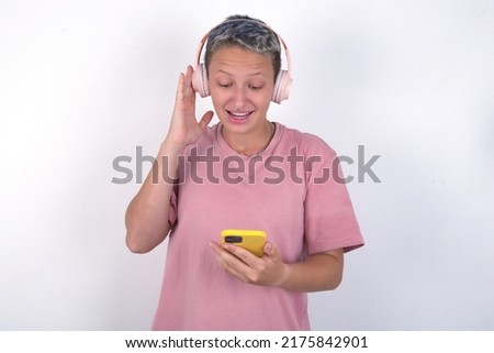 Happy Caucasian woman with short hair wearing pink T-shirt over white wall feels good while focused in screen of smartphone. People, technology, lifestyle