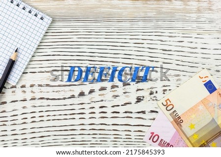 DEFICIT - word (text) and euro money on a white wooden table, notebook, notepad. Business concept: buying, selling, commerce (copy space).