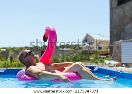 A fat man with a beard and mustache in sunglasses bathes in a framed pool with blue water and a pink inflatable flamingo under a blue sky in a poor area in the slums
