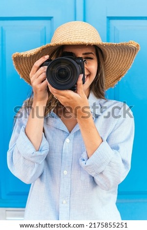 Female traveler in straw hat taking picture on professional photo camera while standing near blue building on street during summer vacation in Lagos in Portugal