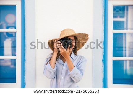 Female traveler in straw hat taking picture on professional photo camera while standing near blue building on street during summer vacation in Lagos in Portugal