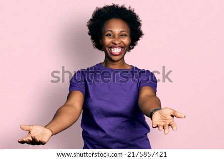 African american woman with afro hair wearing casual purple t shirt smiling cheerful offering hands giving assistance and acceptance. 