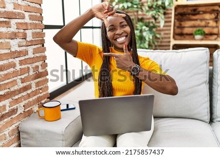 African woman sitting on the sofa using laptop at home smiling making frame with hands and fingers with happy face. creativity and photography concept. 